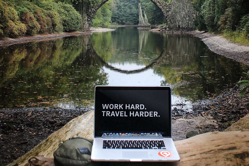A computer with "work hard, travel harder" on its screen with a river in the background